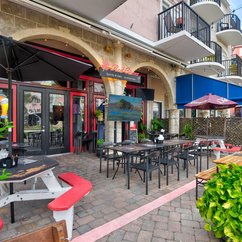 Outdoor patio seating at Red Clover Spirits and Eats cocktail bar, with umbrellas, picnic tables, plants, and historic stone arches.