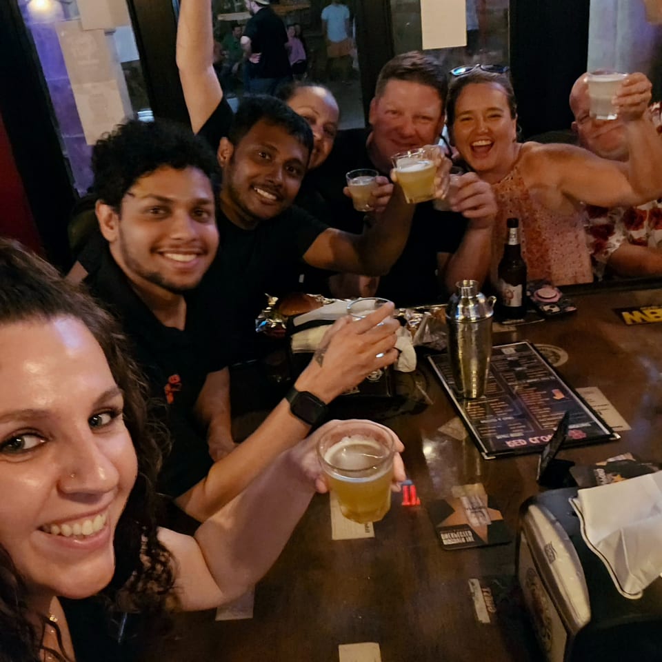 Friends smile and raise drinks together at a lively restaurant bar, with menus, food, and cocktail equipment on the table.