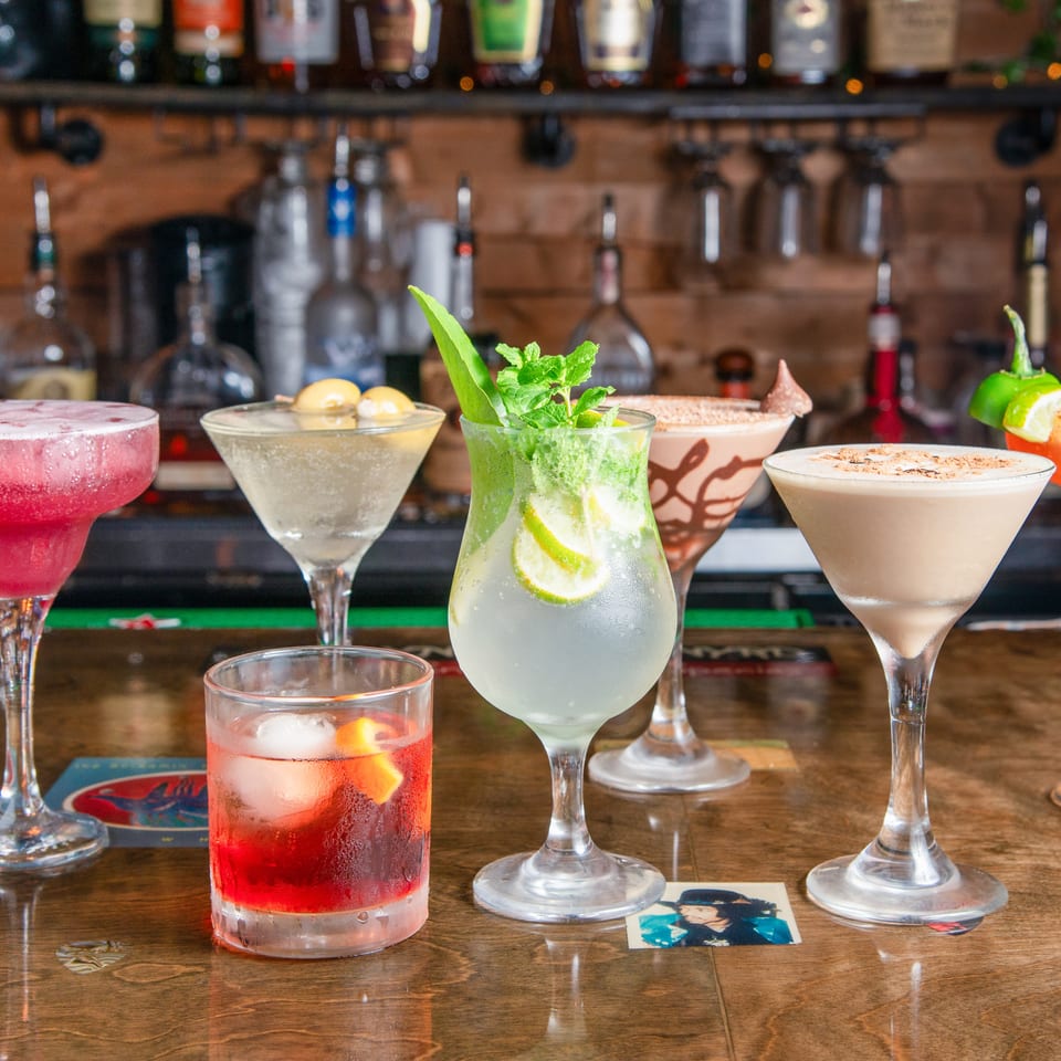 Colorful cocktails, including margaritas, martinis, and mint-garnished drinks, line a wooden bar beneath shelves of liquor bottles.