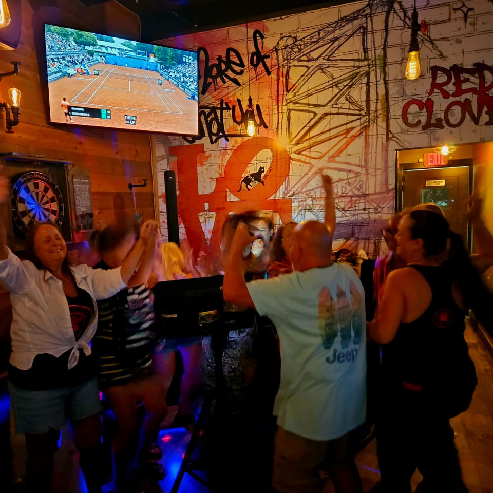 Group of patrons dancing in a dimly lit bar with colorful lights, graffiti-style murals, a dartboard, and televised tennis match.