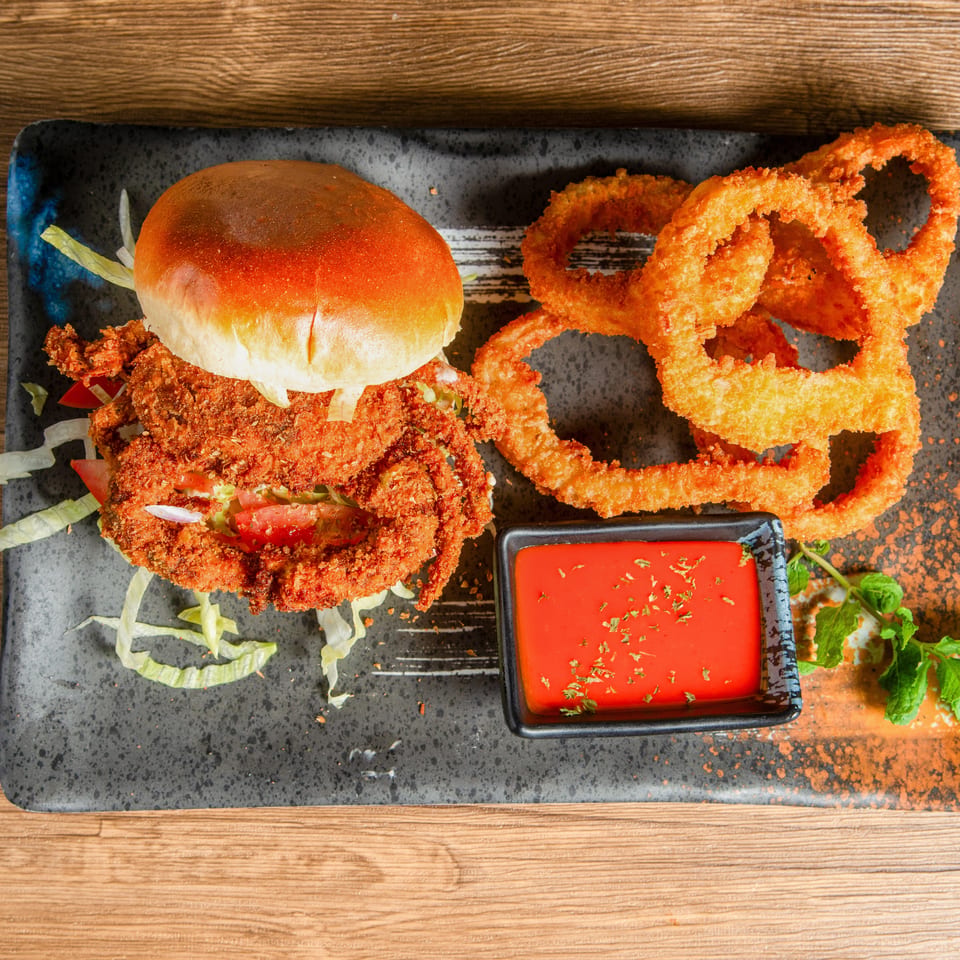 Crispy fried chicken burger with lettuce and tomato, onion rings, and red dipping sauce on a black plate.