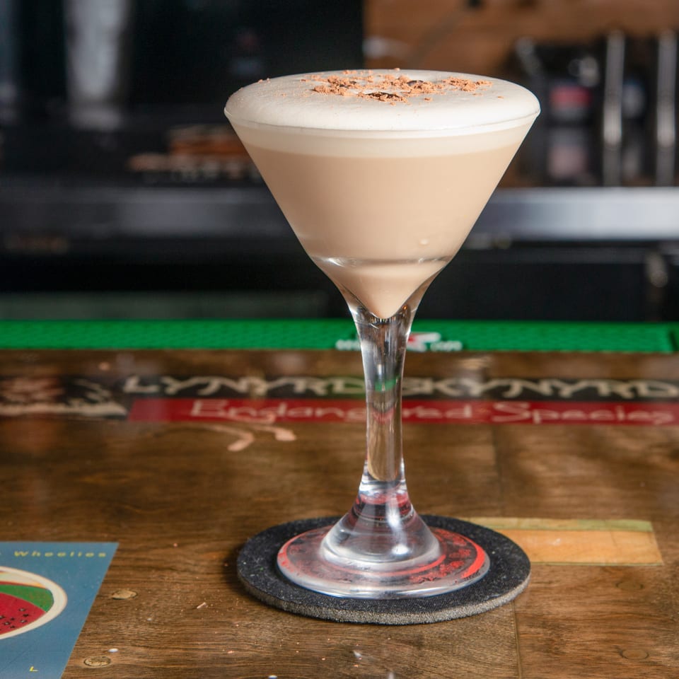 Creamy cocktail topped with grated chocolate served in a martini glass on a wooden bar counter.