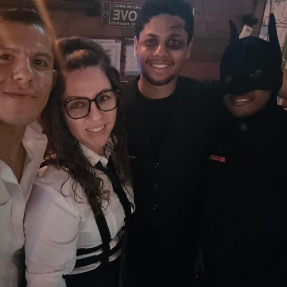 Four people smile for a selfie inside a dimly lit restaurant, including someone wearing a Batman costume.