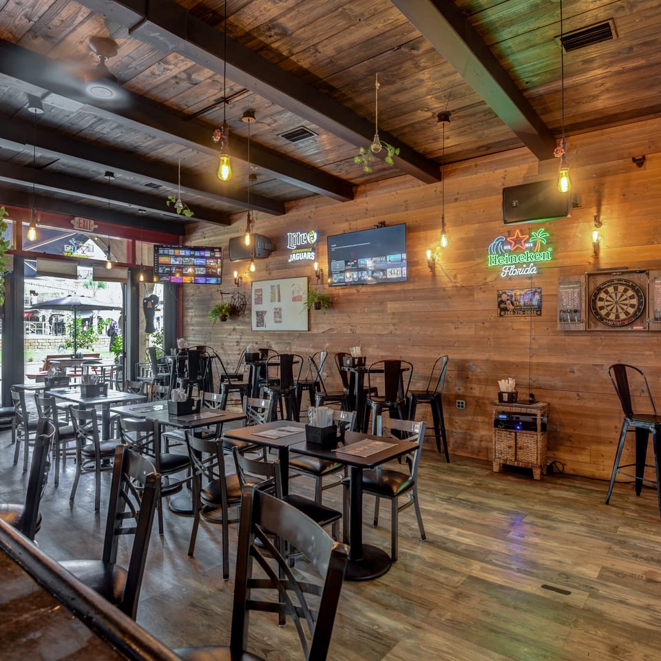 Rustic restaurant interior with wooden walls, pendant lights, dining tables, televisions, a dartboard, and a visible patio entrance.