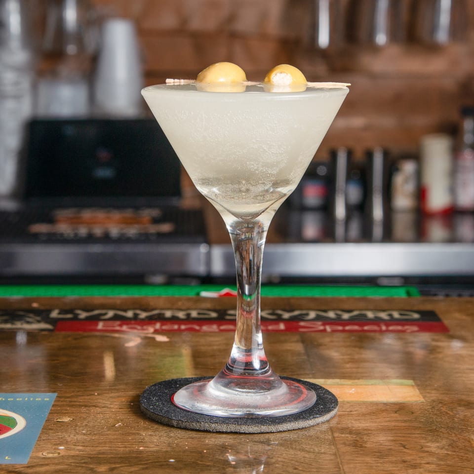 Frosty pale cocktail in a martini glass, garnished with two yellow citrus spheres, sits on a wooden bar.