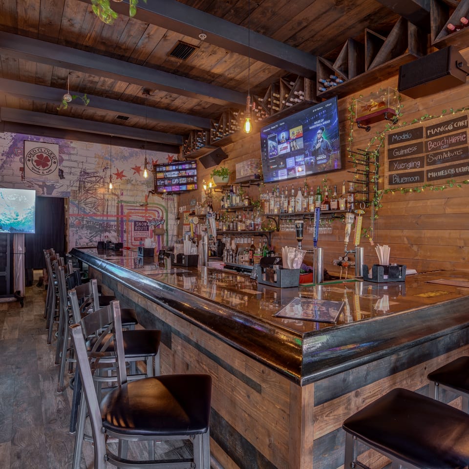 Rustic restaurant bar with polished wood counter, stools, liquor shelves, chalkboard drink menu, televisions, and colorful wall murals.