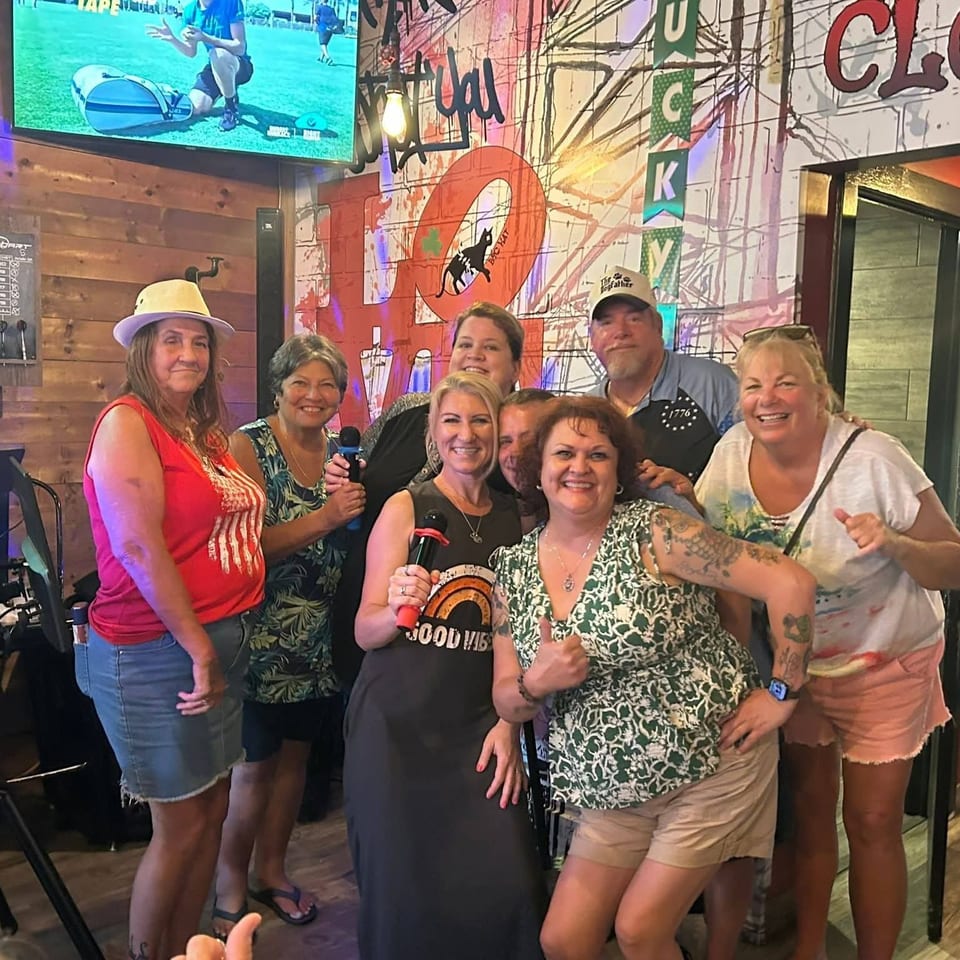 Group of smiling adults pose inside a lively restaurant beside a dartboard, microphone, television, and colorful graffiti-style decor.