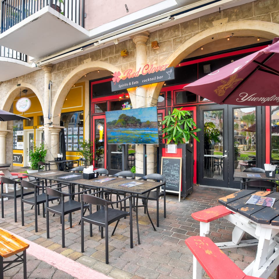 Outdoor patio seating outside Red Clover Spirits & Eats cocktail bar, with umbrellas, picnic tables, and historic stone arches.