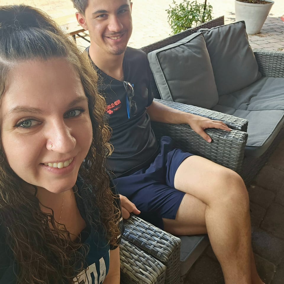 Two people smile for a selfie while seated on wicker patio furniture at an outdoor restaurant.