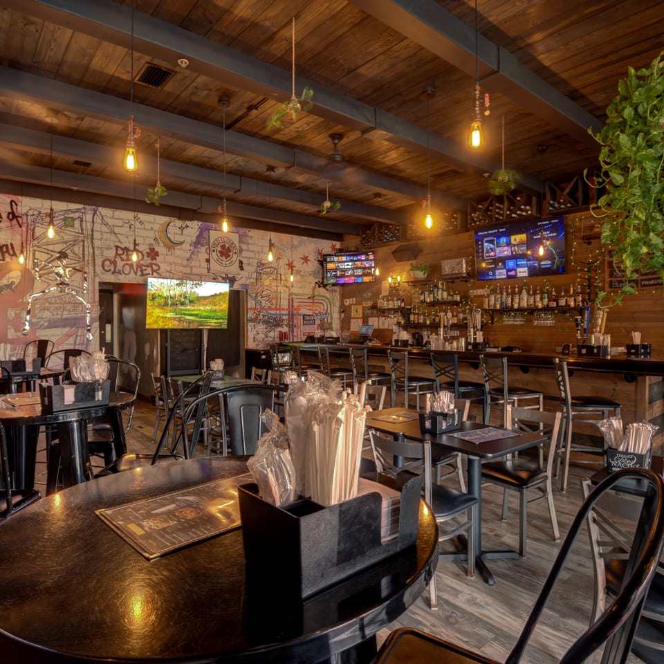 Warmly lit restaurant bar with wooden ceiling, mural walls, televisions, liquor shelves, and neatly arranged tables and chairs.