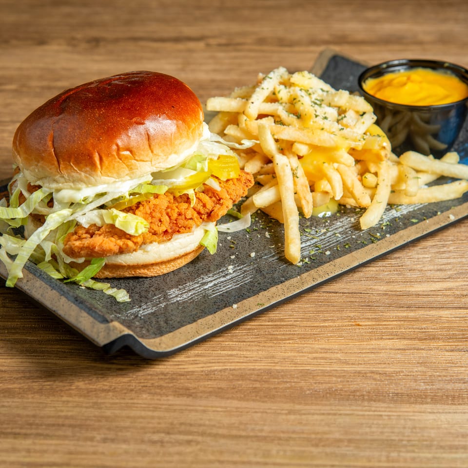 Crispy fried chicken sandwich with lettuce and pickles, seasoned fries, and cheese sauce on a slate platter.