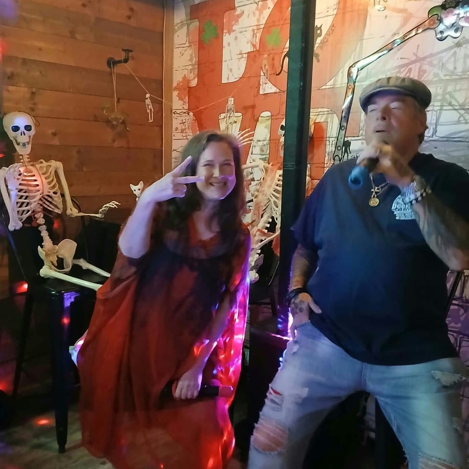 Two people perform in a dimly lit restaurant decorated with skeletons, colorful lighting, and graffiti-style artwork.
