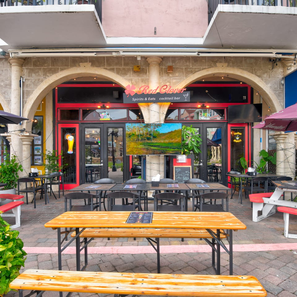 Red Clover Spirits & Eats cocktail bar storefront with arched stone facade, colorful signage, umbrellas, and outdoor tables.