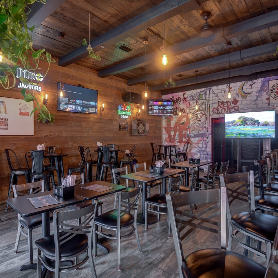 Rustic restaurant dining room with wooden beams, numerous tables and chairs, hanging plants, televisions, and colorful graffiti-style wall art.