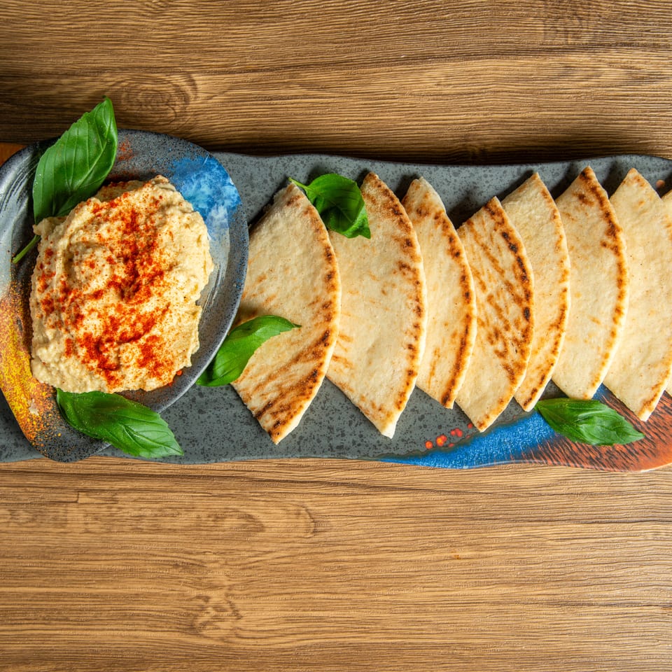 Overhead view of hummus sprinkled with paprika beside grilled pita wedges, basil leaves, and colorful serving platter.