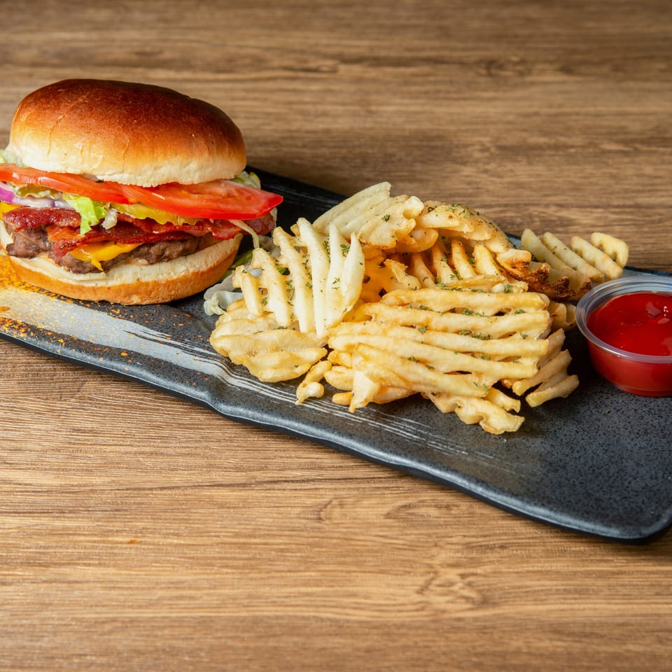 Bacon cheeseburger with lettuce, tomato, onion, and pickles served alongside seasoned waffle fries and ketchup.