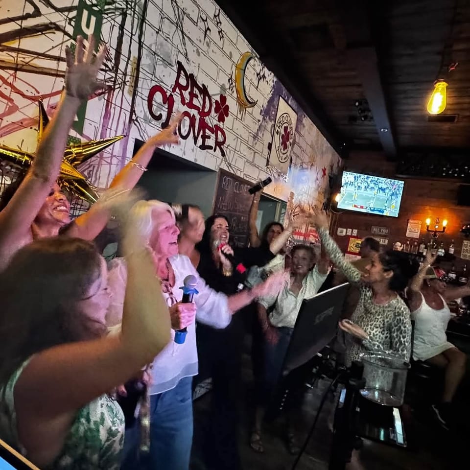 Energetic bar crowd sings and raises hands near a performer, surrounded by televisions, wall art, and celebratory balloons.
