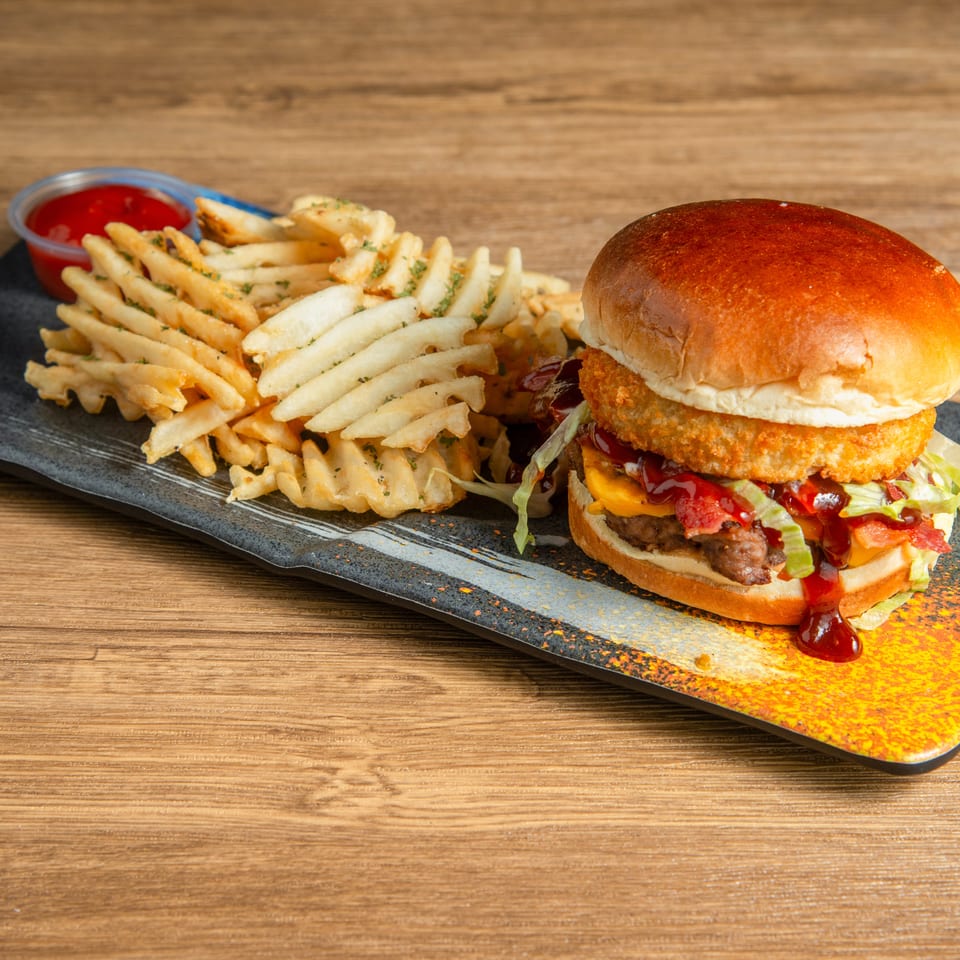 Crispy chicken burger with lettuce, tomato, cheese, and sauce served alongside seasoned waffle fries and ketchup on a black platter.