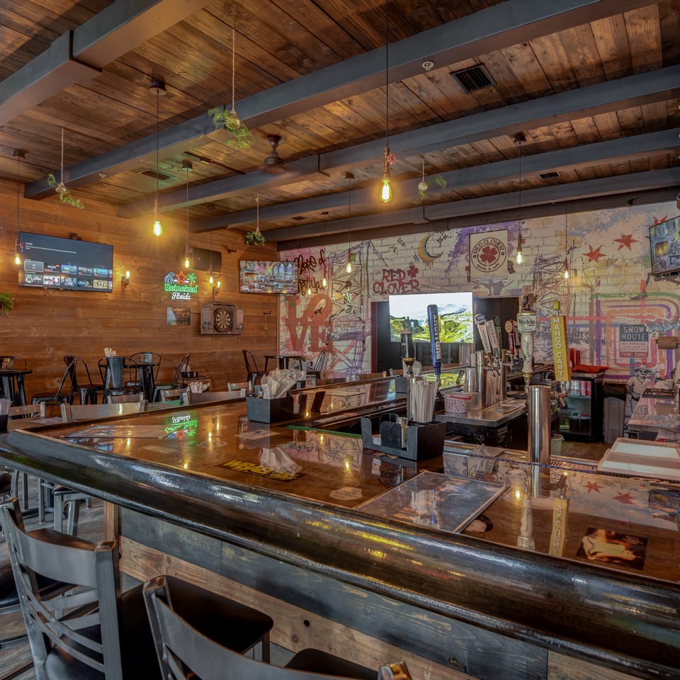 Rustic sports bar interior with a polished counter, bar stools, taps, televisions, and colorful wall murals.