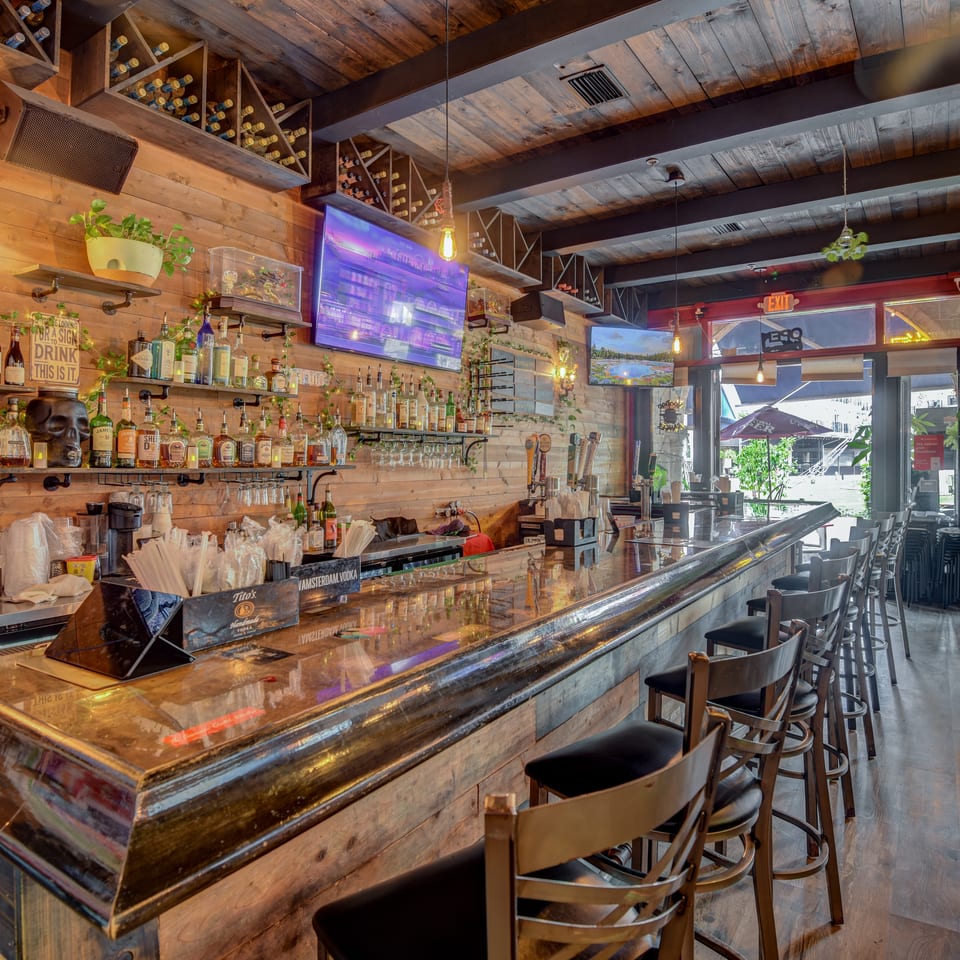 Rustic restaurant bar with wood-paneled walls, liquor shelves, hanging plants, television screens, and rows of barstools.