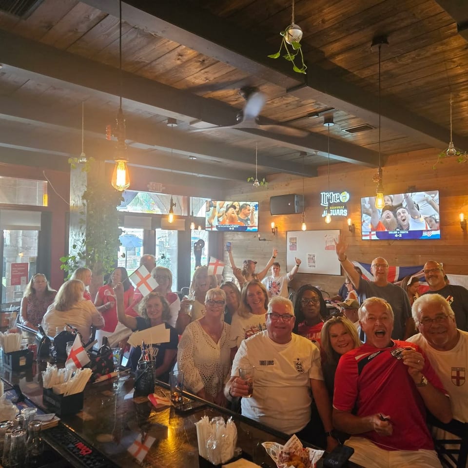Crowded sports bar with cheering fans in soccer jerseys, flags, drinks, and televisions showing a live match.