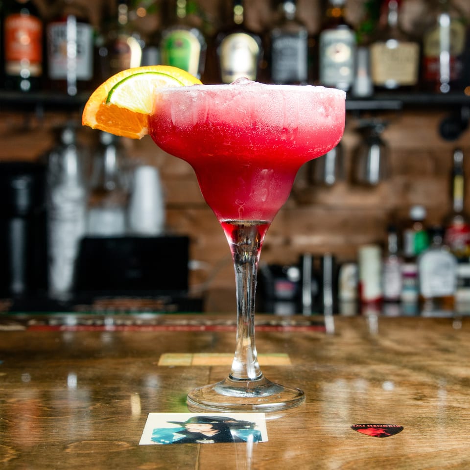 Frosty red margarita in a stemmed glass, garnished with lime and orange wedges on a wooden bar.
